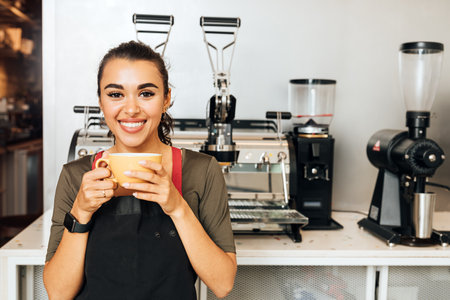 Beautiful cheerful barista holding a mug and looking at camera. Smiling waitress taking a break during a working day.の写真素材