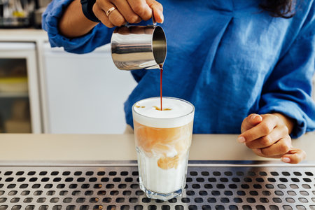 Cropped shot of female barista making a glass of latte while standing behind counter. Hands of woman pouring coffee into a cup milk.の写真素材
