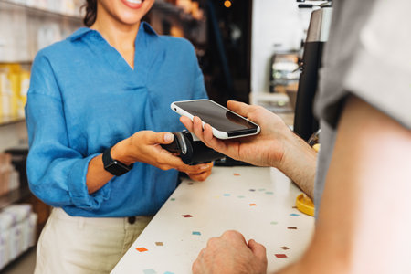 Hand of a customer at a cafe paying his bill using a mobile phone. Female entrepreneur holding a POS...