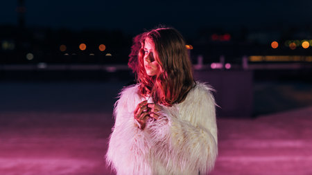 Young blonde woman standing on a roof in front of a pink neon light and looking awayの写真素材