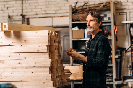 Senior man standing near a bunch wooden parts of furniture in a workshop and looking awayの写真素材