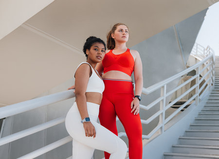 Two plus size females in sportswear standing on a staircase. Young women relaxing after exercises outdoors.の写真素材