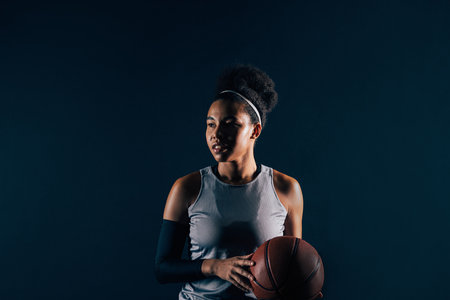 Female basketball player in sportswear over black backdrop. Young professional basketball player with ball, wearing team attire.の写真素材