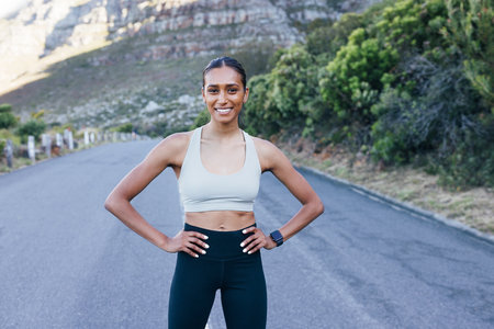 Smiling confident sportswoman standing on road with hands on hips and looking at camera