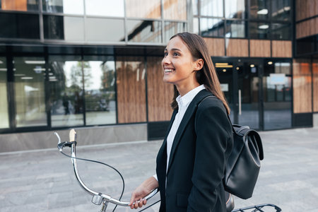 Happy female with bicycle and backpack walking outside. Young smiling businesswoman with a bicycle walking on the street.の写真素材
