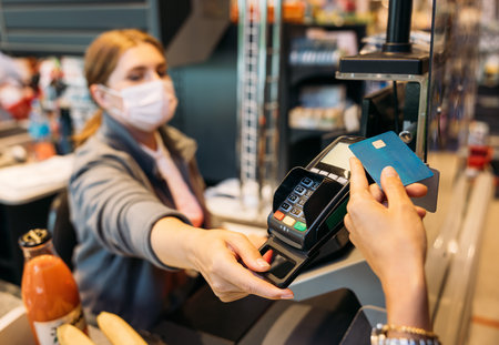 Unrecognizable woman's hand holding a credit card with NFC chip while the cashier in a mask turns...