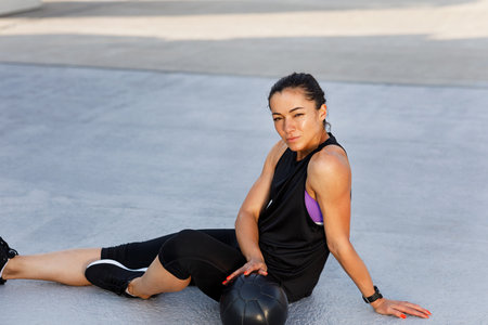 Young sportswoman relaxing during workout. Female in sportswear sitting with medicine ball.