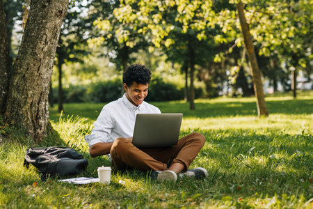 Happy male college student typing on laptop while sitting on a grass in parkの写真素材