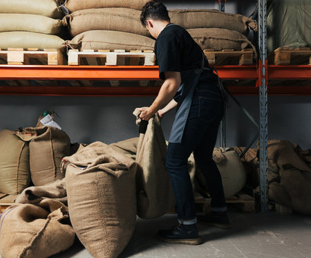 Back view of a young warehouse worker moving a bag. Barista is managing bags of coffee next to the...