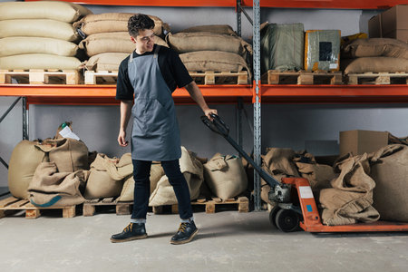 Young blue-collar worker is pulling a manual pallet jack at a warehouse. Male working at a coffee shop warehouse.の写真素材