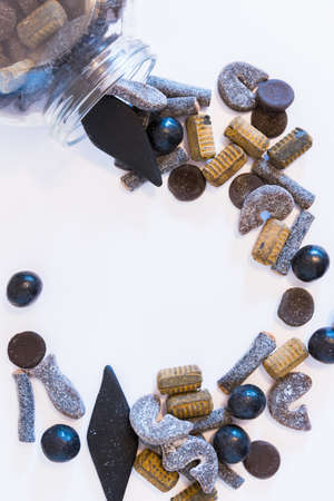 Flat-lay arrangement of variety of double salted licorice spilling from glass candy jar onto white...