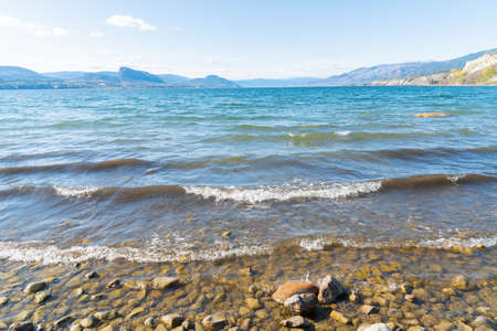 Close-up view of scenic Okanagan Lake reflecting blue sky and sunshine, with view of beach and mountainsの写真素材