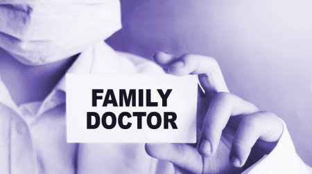 A doctor in face mask and medical clothing shows a card with the text FAMILY DOCTOR. Medical concept.の写真素材