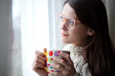 Beautiful young woman sitting by the window having hot drink