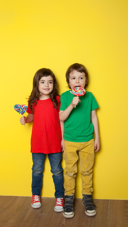 Adorable boy and girl having great time while licking lollipops, on red and yellow backgroundの写真素材