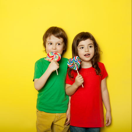 Adorable boy and girl having great time while licking lollipops, on red and yellow backgroundの写真素材