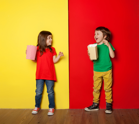 Adorable boy and girl having great time eating popcorn, on red and yellow backgroundの写真素材