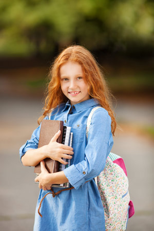 Adorable little red-haired girl posing with books and backpack