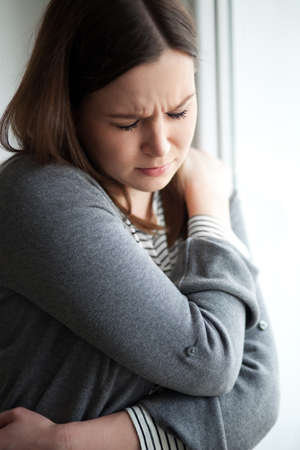 Sad looking young woman standing by the window, on white backgroundの写真素材