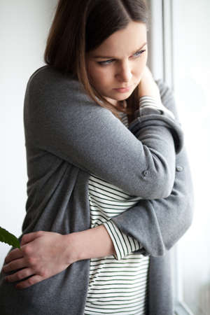 Sad looking young woman standing by the window, on white backgroundの写真素材