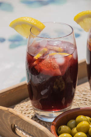 Close up of a typical spanish drinking glass of red wine with soda, ice cubes and slices of lemon next to a plate with olives on a wooden tray. Sangria concept.の写真素材