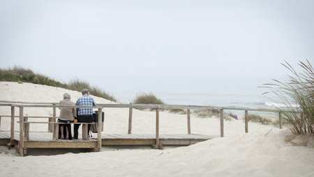 Costa Nova, Portugal - August 2018: Cute old couple sitting in wooden bench looking at sea in Costa Nova beach.のeditorial素材