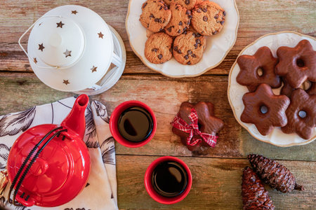 Beautiful rustic wooden table full of cookies and tea. Red teapot with full teacups, pine cones, chocolate chip cookies.の写真素材