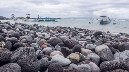 Horizontal photo of a stone beach and traditional Balinese fishing boats and gazebos in the distance in pantai Candidasa beachfront, during a cloudy day. Bali, Indonesiaの写真素材