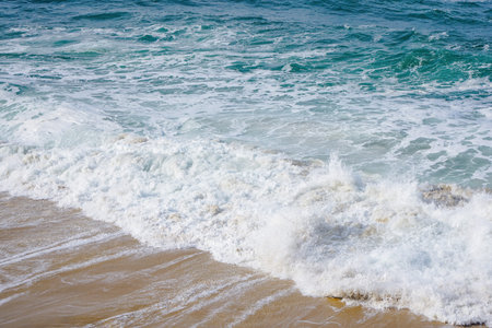 Horizontal photo of turquoise ocean water with small white foam waves gently crashing into golden...