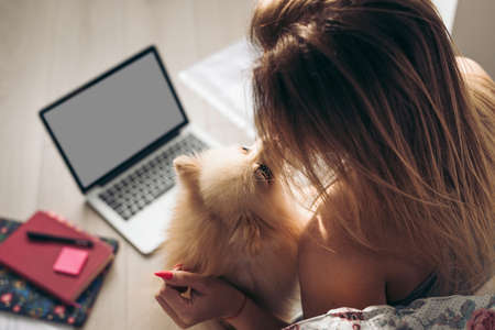 Beautiful woman works from home with dokuments and laptop. The dog helps her. Girl kisses her pomeranian spitz. The concept of working from home. Concept Love Care, Comfort and Companionship. WFHの写真素材