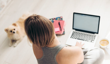 Work from home and pets. Pomeranian spitz and a young woman working with a laptop on a wooden floor.
