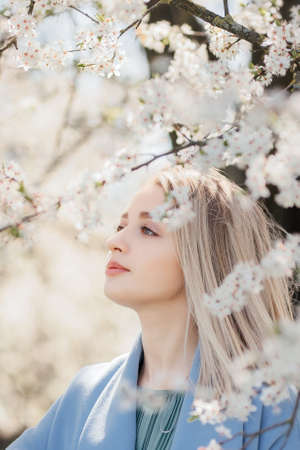 Stylish beautiful woman sensually posing among blooming apple branches in sunny spring, calm moment. Portrait of happy young blonde female in blue jacket embracing in white flowersの写真素材
