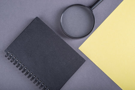 Notebook with a magnifying glass and yellow paper on a gray backgroundの写真素材