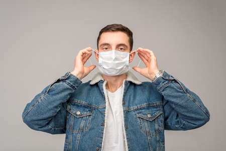 Against a light background, a man in a denim jacket puts on a medical mask. Virus protection. Studio photo.の写真素材