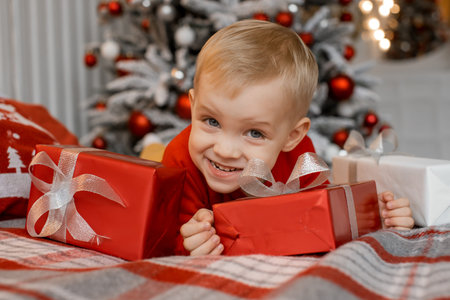 Beautiful little blond boy with a playful smile lying on bed with red Christmas presents and...