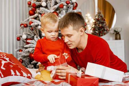 Happy family dad and son in red sweaters hold Christmas gifts next to the New Year Tree at home. Christmas time.の写真素材