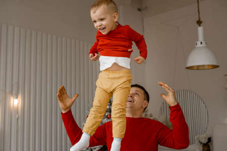 Cheerful happy father throwing up his adorable laughing son in bright white room with Christmas interior at home. Family, holiday, parenthood, playing, game, childhood and leisure time conceptの写真素材
