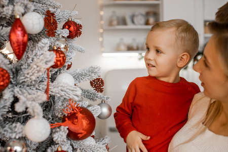 Charming mother in white sweater holding her little son next to beautifully decorated Christmas tree in the houseの写真素材