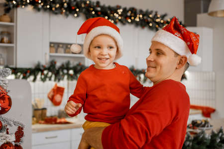Adorable cute little boy sitting on his father hands in the kitchen. Both are in festive santa hats and red sweaters.の写真素材