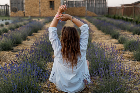 female fashion. Back view of young woman sitting among lavender flowers in summer sunset wearing white shirt.の写真素材