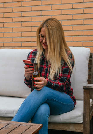Young blonde girl watching mobile, smiling and having a beer on a terraceの写真素材