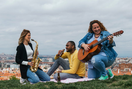 Happy interracial friends sitting in a park playing saxophone and guitar and laughing