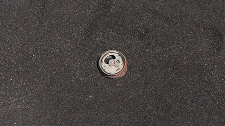 Rusty aluminium beverage can washed up on a dark sand beach, impacting the environment and polluting...