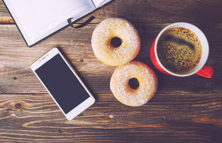 Donuts and coffee lying on rustic wooden background with open notepad and mobile phone. Top view. Retro toned picture.の写真素材