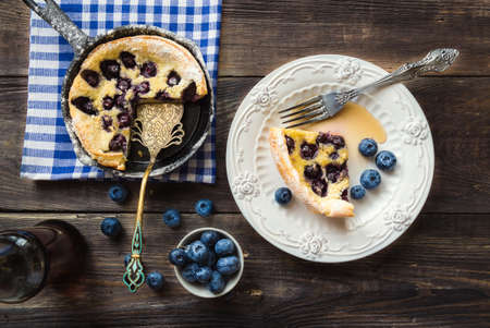Piece of fresh homemade Dutch Baby pancake with blueberries on the plate with fork, skillet, bowl with berries and a bottle with maple syrup on rustic wooden background. Top view.の写真素材