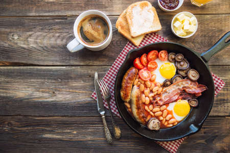 Fried eggs, sausages, bacon, beans and mushrooms in iron skillet, toasts, coffee, butter and jam on rustic woodenの写真素材