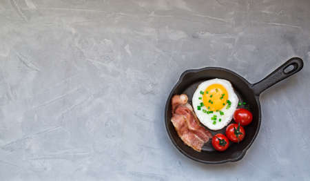 Fried egg with bacon and cherry tomatoes in iron skillet on light gray concreteの写真素材