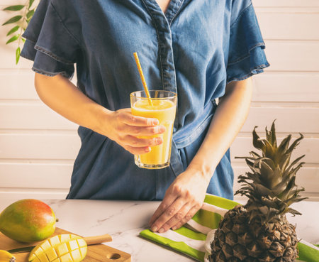 Young woman in denim dress holding a glass with mango and pineapple smoothie in her hand. Healthy...