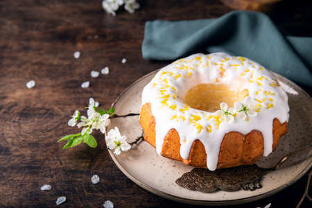Fresh homemade lemon bundt cake decorated with white glaze and zest on rustic wooden background with branches of blossoming plum.の写真素材