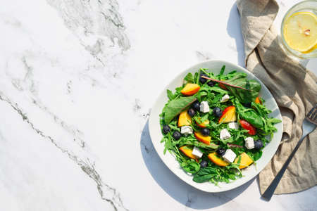 Fresh homemade salad with nectarines, blueberries, arugula, spinach and feta cheese on white marble background with hard shadows. Healthy vegetarian food. Top view. Space for text.の写真素材